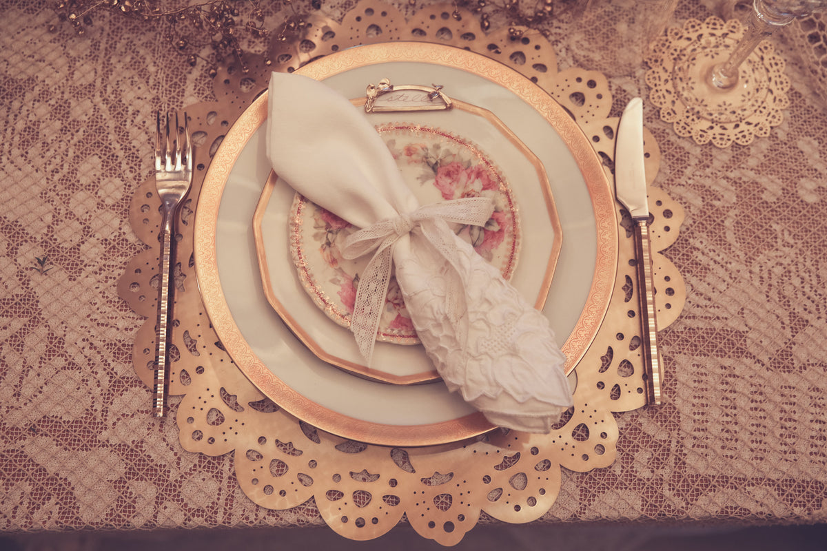 Gold Eyelet Doily Charger under gilded vintage china and a lace-tied napkin on a lace tablecloth, with an Eyelet Doily Coaster - Ariana Ost