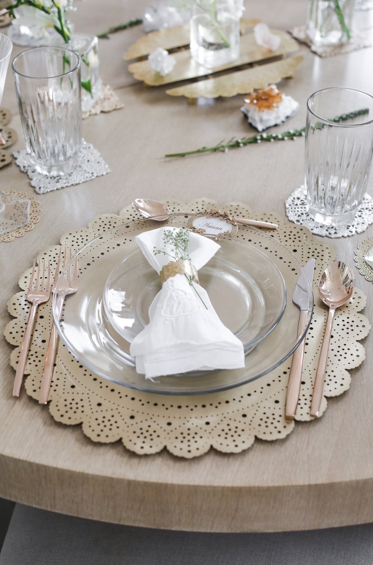 Gold Lace Doily Charger under a glass plate with a lace doily napkin ring, white linen and rose gold flatware on a pale wood table - Ariana Ost