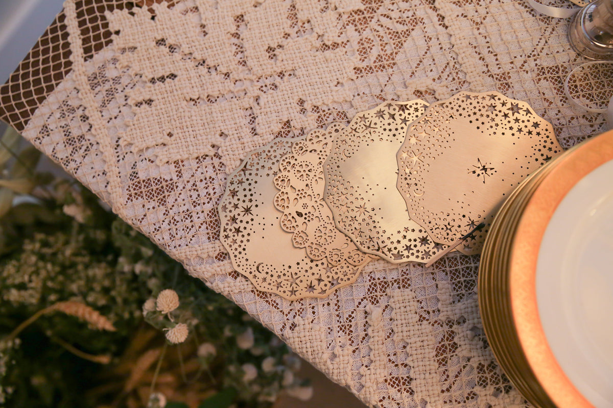Gold Celestial Coasters fanned out with a Lace Doily Coaster on a vintage lace tablecloth beside gilded china - Ariana Ost