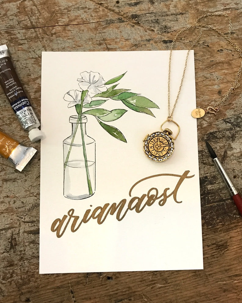 Clock Locket necklace on a hand-lettered Ariana Ost watercolor card, with paint tubes and a brush on a rustic wood table - Ariana Ost
