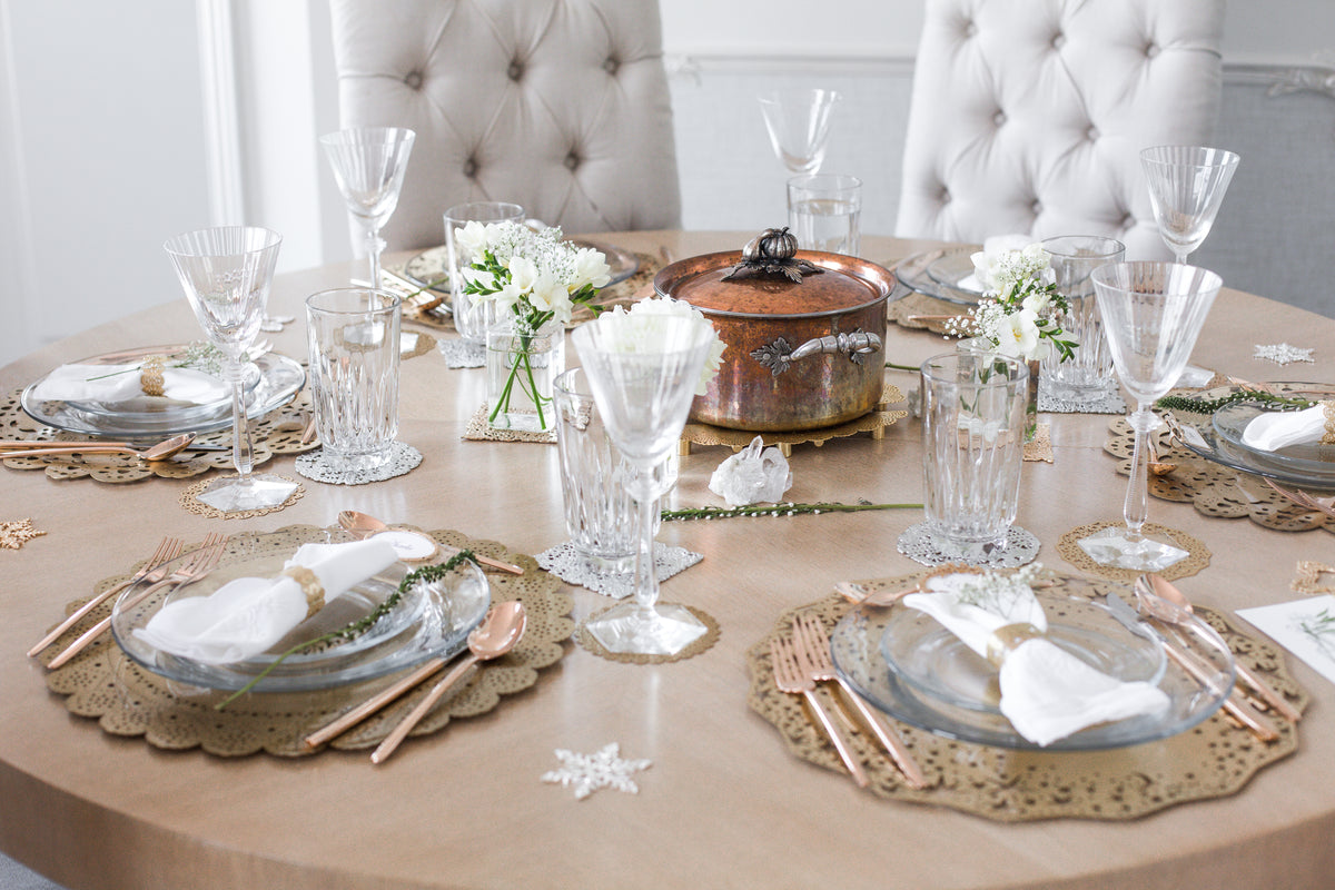 Round dinner table set with gold laser-cut doily chargers, coasters and napkin rings, crystal glasses and white flowers in a white Parisian-style dining room - Ariana Ost