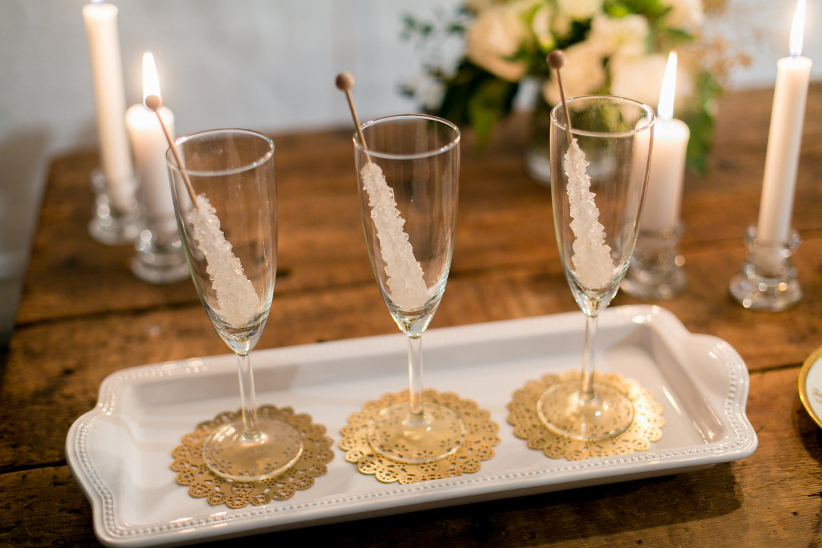 Gold Eyelet Doily Coasters under champagne flutes with rock candy stirrers at a holiday party - Ariana Ost